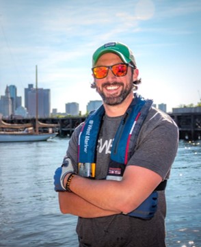 Photo of Adam on a sunny day by the water. He is wearing a life jacket and sunglasses, and is smiling at the camera. 