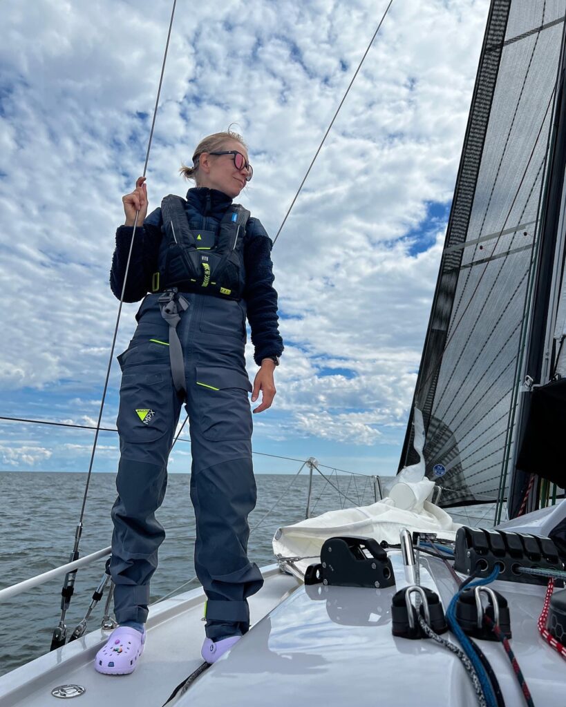 Noora standing on the bow in foul weather gear and sunglasses, holding a line and looking off to the starboard. 